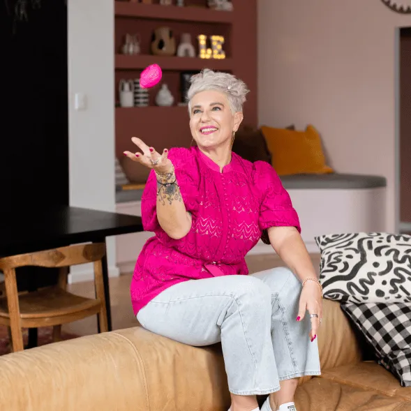 Dr Leanne Elich, in a bright pink top and light jeans, sits on a sofa playfully tossing a pink object in a modern living room.